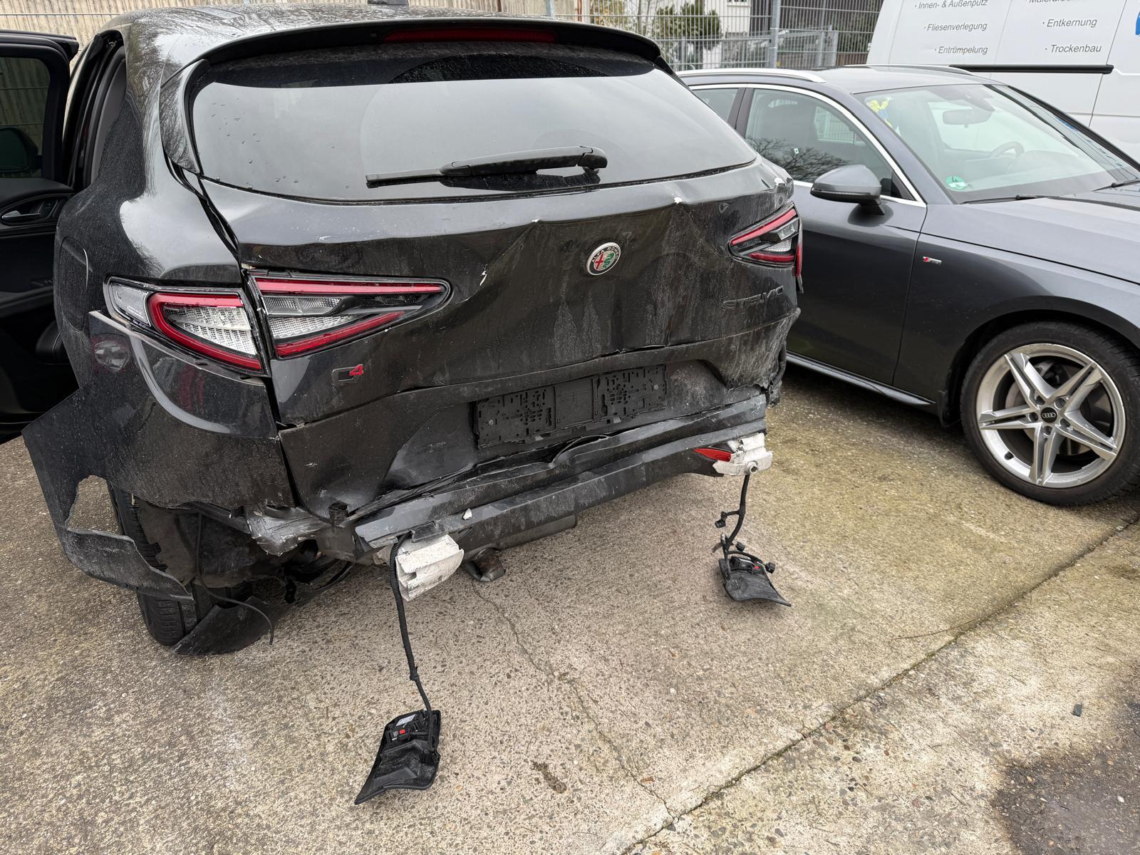 Heckschaden: Stark beschädigte Rückseite eines schwarzen Autos mit herabhängenden Kabeln.